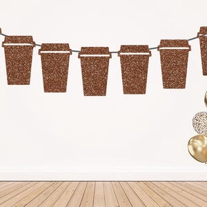 May include: A decorative banner featuring seven brown glittery coffee cup cutouts strung together on a thin cord, accompanied by a cluster of gold and glittery gold balloons positioned at the bottom right corner of the display.