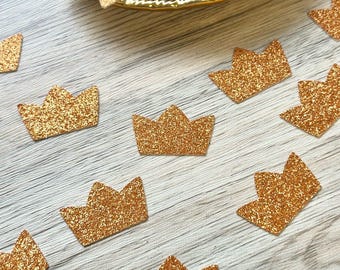 May include: A collection of small, flat, crown-shaped cutouts with a textured, shimmering gold glitter finish. Several individual crown pieces are scattered across the surface, while a larger cluster is contained within a gold-toned dish.