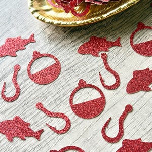 May include: A collection of red glitter paper cutouts shaped like fish, fishing hooks, and round bobbers. The cutouts are scattered across the surface, with a gold-coloured bowl filled with additional red glitter shapes visible at the top.