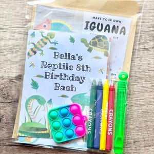 May include: A clear plastic party favor bag containing a Make Your Own Iguana paper craft kit, a card labeled Bella's Reptile 8th Birthday Bash with reptile illustrations, four wax crayons in green, blue, yellow, and red, and a green bubble wand.