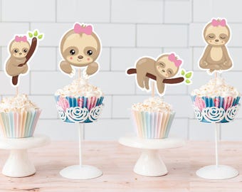 May include: Four cupcakes decorated with sloth themed toppers. The cupcakes are on white cake stands. The sloths are wearing pink bows and are in various poses, including sleeping, sitting, and hanging from a branch.