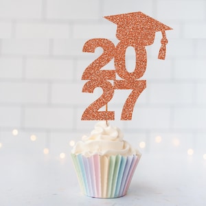 May include: A copper-colored glitter cupcake topper featuring the numbers 2027 and a graduation cap icon. The topper is mounted on a thin wooden pick and is inserted into a cupcake with a pastel-striped paper liner, creating a festive display.