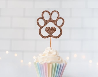 May include: A brown glittery paw print cupcake topper with a central heart cutout, attached to a thin wooden pick and inserted into a cupcake with a white frosting swirl and a pastel-colored pleated paper liner, creating a decorative accent.