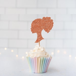 May include: A cupcake with a light-colored frosting and a decorative topper. The topper is a silhouette of a woman's profile with a bun, covered in copper glitter. The cupcake is in a pastel-colored paper cup.