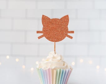 May include: A copper-colored glitter cat-shaped cupcake topper on a thin wooden stick, inserted into a cupcake with white frosting and a pastel-striped paper liner. The cat silhouette features pointed ears and horizontal whiskers on each side.