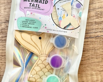 May include: A paint your own mermaid tail wood ornament craft kit in a clear pouch. The kit includes a wooden mermaid tail cutout, a paint brush, three small paint pots in blue, purple, and green, and an ornament cord. The label reads Paint Your Own Mermaid Tail Wood Ornament and Little Bella Boutique.