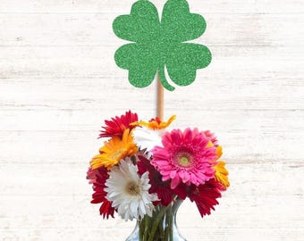 May include: A green glittery four-leaf clover cutout attached to a wooden stick, placed inside a clear glass vase filled with a vibrant bouquet of multi-colored gerbera daisies in shades of pink, white, orange, and deep red.