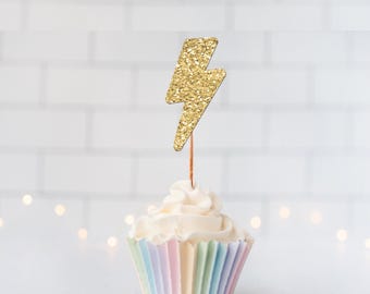 May include: A cupcake topper featuring a gold glitter lightning bolt shape attached to a thin wooden pick. The cupcake is topped with white frosting and sits in a pleated paper liner with vertical stripes in pastel shades of pink, blue, and yellow.