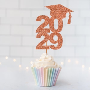 May include: A copper-colored glitter cupcake topper featuring the numbers 2029 topped with a graduation cap. The topper is inserted into a white frosted cupcake with a pastel-striped paper liner, creating a festive display for a graduation event.