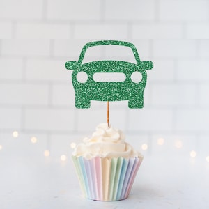 May include: A green glitter car-shaped cupcake topper attached to a thin wooden pick, positioned vertically in the center of a white frosted cupcake with a pastel-colored pleated paper liner, featuring a simple silhouette of a vehicle.