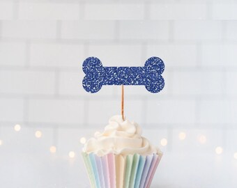 May include: A cupcake topper shaped like a dog bone with a dark blue glitter finish, attached to a thin wooden pick. The topper is inserted into a cupcake with white frosting and a pleated paper liner featuring vertical stripes in pastel colors.