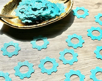 May include: A collection of small, turquoise gear-shaped confetti pieces with a glittery texture. Some pieces are scattered individually, while a larger pile is contained within a gold-colored, leaf-shaped decorative dish.