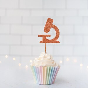 May include: A cupcake with a light-colored frosting and a pastel-colored liner. A copper-colored glitter microscope cake topper is inserted into the cupcake. The background is a white brick wall with soft, out-of-focus lights.