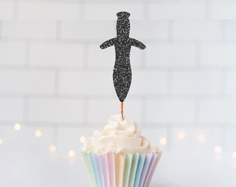 May include: A black glittery dagger-shaped cupcake topper is inserted into a white frosted cupcake with a pastel-colored pleated paper liner. The dagger silhouette features a pointed blade and a crossguard, mounted on a thin wooden pick.