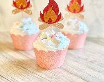 May include: Three cupcakes in glittery peach-colored wrappers topped with white frosting, colorful sprinkles, and a glittery red, orange, and gold campfire-shaped paper topper attached to a small wooden pick inserted into the center.