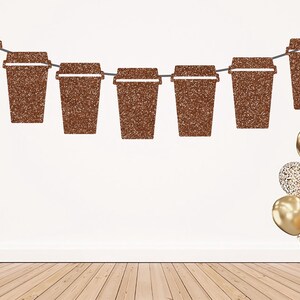 May include: A decorative banner featuring seven brown glittery coffee cup cutouts strung together on a thin cord. To the right, a cluster of gold balloons includes solid metallic spheres and glitter-covered spheres, all tied together at the base.
