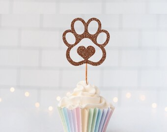 May include: A brown glittery paw print cupcake topper with a small heart cutout in the center, attached to a thin wooden pick and inserted into a cupcake with a pastel-colored paper liner featuring vertical stripes of light blue, pink, and yellow.