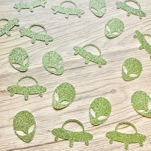 May include: A collection of green glitter confetti pieces shaped like alien heads and flying saucers. The individual cutouts feature a sparkling texture and are scattered across the frame, showcasing the distinct silhouettes of extraterrestrial beings.
