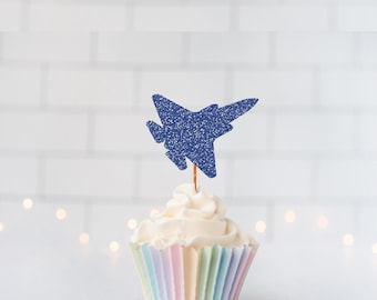 May include: A cupcake topper shaped like a fighter jet, featuring a dark blue glitter finish. The jet is attached to a thin wooden pick and is positioned upright in the center of a cupcake with white frosting and a pastel striped paper liner.