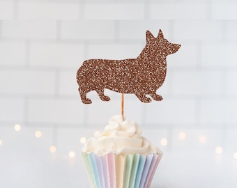 May include: A brown glittery Corgi dog silhouette cake topper attached to a thin wooden pick, inserted into a cupcake with white frosting in a pastel-striped paper liner. The dog shape is detailed with a textured, sparkling finish.