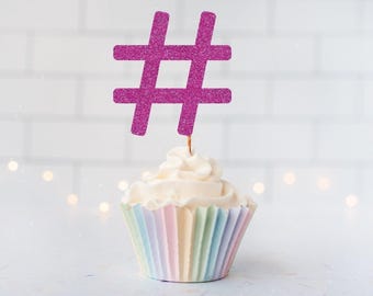 May include: A cupcake topper featuring a magenta glittery hashtag symbol attached to a thin wire stem. The cupcake is topped with white swirled frosting and sits in a pleated paper liner with alternating pastel stripes of pink, yellow, and blue.