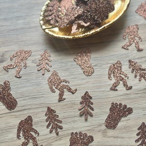 May include: A collection of copper-colored glittery confetti shapes including silhouettes of Bigfoot, pine trees, and footprints. Some pieces are contained within a small, oval-shaped gold-toned dish, while others are scattered across the surface.