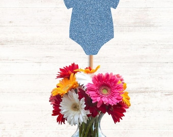 May include: A blue glittery baby bodysuit cutout attached to a wooden stick, positioned above a clear glass vase filled with a vibrant bouquet of pink, white, orange, and red gerbera daisies and other assorted flowers.