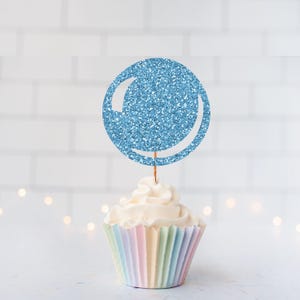 May include: A light blue glitter cupcake topper shaped like a circle with a crescent cut-out, on a cupcake with a pastel rainbow wrapper. The cupcake has white frosting.