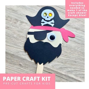 May include: A paper craft kit featuring a pirate face with a black hat, white skull and crossbones with yellow rhinestone eyes, a pink headband, a black eye patch, a single googly eye, and a black beard, all attached to a wooden craft stick.