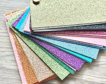 May include: A fan of colorful glitter paper swatches in various colors, including gold, pink, green, purple, and blue. The swatches are held together with a small, round, beige button. The glitter paper has a textured surface.