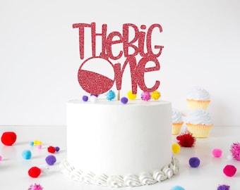 May include: A red glitter cake topper featuring the text "The BIG One" with a circular graphic element, positioned on top of a plain white frosted cake decorated with a piped border and surrounded by small colorful pom-poms on the surface.