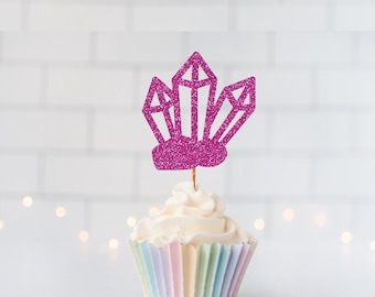 May include: A bright magenta glitter cupcake topper shaped like a cluster of three crystals, mounted on a thin wooden pick and inserted into a white frosted cupcake with a pastel-colored paper liner featuring vertical stripes of pink, blue, and yellow.