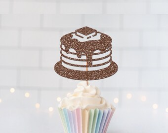 May include: A brown glittery pancake-shaped cake topper featuring a stack of pancakes with syrup and a pat of butter on top, attached to a thin wooden pick, inserted into a white frosted cupcake with a pastel-colored paper liner.