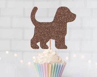 May include: A brown glittery dog-shaped cupcake topper attached to a thin wooden pick, positioned vertically above a single cupcake with white frosting in a pastel-striped paper liner. The dog silhouette features a curved tail and rounded ears.