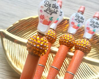 May include: A set of orange glitter pens featuring a topper shaped like a lightbulb with the text Be the Light and floral accents. Each pen includes a large, round, amber-colored rhinestone bead above the barrel and a matching orange glitter cap.