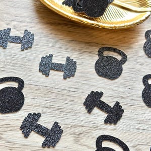 May include: A collection of black glittery paper cutouts shaped like dumbbells and kettlebells. Some of the fitness-themed confetti pieces are scattered across the surface, while others are gathered inside a gold-colored leaf-shaped dish.
