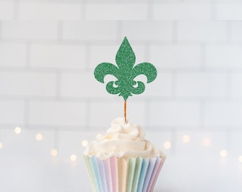 May include: A green glitter fleur-de-lis cupcake topper on a thin wooden pick, inserted into a cupcake with white frosting and a pastel-colored pleated paper liner. The topper features a classic three-petal design with a textured, sparkling finish.