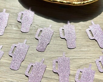 May include: A collection of small, light purple glittery paper cutouts shaped like tumblers with handles and straws. Some of the cutouts are scattered across the surface, while others are contained within a gold-colored, leaf-shaped dish.
