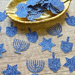 May include: A collection of blue glitter paper cutouts in the shapes of dreidels, menorahs, and six-pointed stars. Some of the cutouts are scattered across the surface, while others are contained within a gold-coloured leaf-shaped dish.