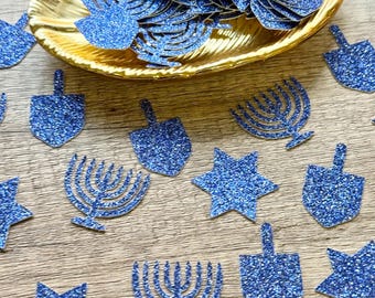 May include: A collection of blue glitter paper cutouts in the shapes of dreidels, menorahs, and six-pointed stars. Some of the cutouts are scattered across the surface, while others are contained within a gold-colored leaf-shaped dish.