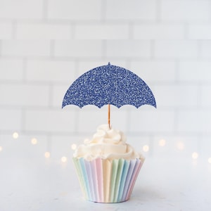May include: A cupcake topped with a blue glitter umbrella cake topper. The cupcake features white frosting and sits in a pastel-coloured pleated paper liner. The umbrella topper is attached to a thin wooden pick inserted into the centre of the frosting.