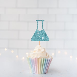 May include: A cupcake with pastel-colored paper and white frosting, topped with a glittery blue beaker-shaped decoration. The cupcake is centered against a white brick wall background with soft, out-of-focus lights.