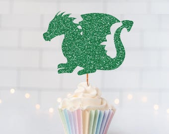 May include: A green glitter dragon cupcake topper attached to a thin wooden pick, positioned vertically on top of a single cupcake with white frosting in a pastel-colored pleated paper liner, showcasing a sparkling texture and silhouette.