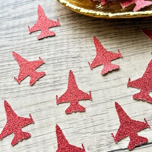 May include: Red glitter airplane-shaped confetti scattered on a wooden surface, with some pieces in a gold-colored dish. The confetti has a sparkly texture and is designed in the shape of a jet plane. Ideal for crafting or party decorations.