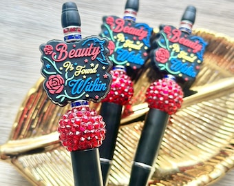 May include: Three black pens featuring a decorative topper with the text Beauty Is Found Within, accented by red roses and small stars. Each pen includes a textured red bead and a silver and blue ring detail near the top of the black barrel.