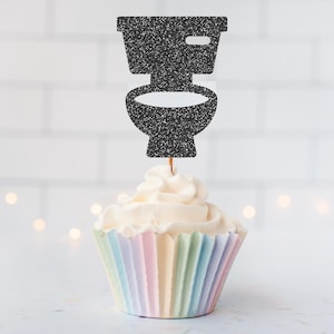 May include: A black glittery toilet-shaped cupcake topper is inserted into a vanilla cupcake with white frosting. The cupcake sits in a pleated paper liner featuring a pastel rainbow gradient of light pink, yellow, blue, and green vertical stripes.