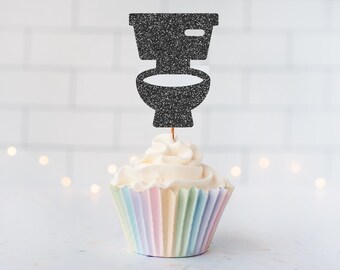 May include: A black glittery toilet-shaped cupcake topper is inserted into a vanilla cupcake with white frosting. The cupcake sits in a pleated paper liner featuring a pastel rainbow gradient of light pink, yellow, blue, and green vertical stripes.