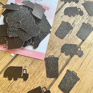 May include: A collection of black glittery die-cut paper shapes featuring silhouettes of garbage trucks and trash cans. The shapes are arranged in a pile on a branded white card and scattered individually, displaying a textured, sparkling finish.