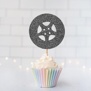 May include: A black glittery circular cupcake topper featuring a five-spoke wheel cutout design. The topper is attached to a thin wooden pick and is positioned vertically in the centre of a white frosted cupcake with a pastel striped paper liner.
