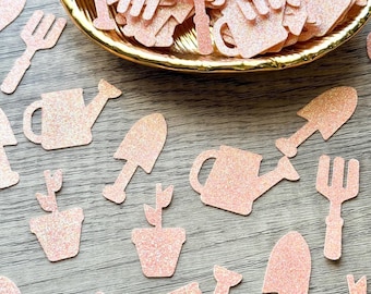 May include: A collection of light pink glittery paper cutouts shaped like gardening tools, including watering cans, shovels, rakes, and potted plants, with a large pile contained in a gold-toned, leaf-shaped decorative dish.